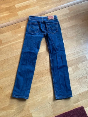 Blå raka jeans från Levi's - Säljer ett par klassiska blå jeans från Levi's med raka ben och femficksmodell. Jeansen har tydliga kontrastsömmar och en ikonisk läderpatch bak i midjan. Tillverkade i slitstark denim med normal midja och knappgylf.pris kan diskuteras. 