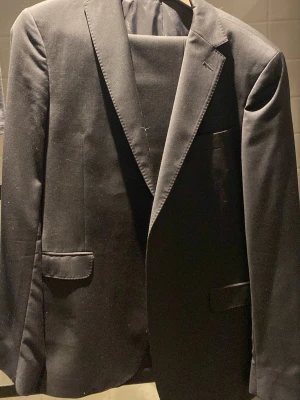 Svart kavaj från Oscar Jacobson - Klassisk svart kavaj från Oscar Jacobson med två framfickor och bröstficka. Kavajen har slimmad passform, elegant slag och är helfodrad med diskret mönstrat innerfoder. Perfekt för dig som vill ha en stilren och tidlös look.