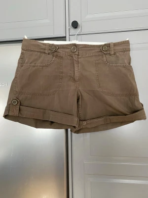 Bruna shorts dam strl 42 - Snygga bruna shorts med uppvikta ben och dekorativa knappar i midjan och vid benslut. Shortsen har två framfickor och är tillverkade i ett mjukt bomullsmaterial. Perfekta för varma dagar och enkel att matcha med olika toppar.