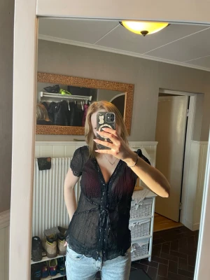 Svart transparent blus med knyt - Snygg svart transparent blus med korta ärmar och knyt i midjan. Djup v-ringning och lätt figurnära passform. Tunn mesh-liknande material som ger en luftig känsla. Perfekt att styla med jeans eller kjol.