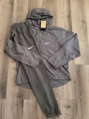 🩶grå Nike repel miller sät med huva🩶 - Snyggt grått träningsset från Nike med jacka och byxor. Jackan har huva, dragkedja och två fickor, medan byxorna har resår i midjan och vid benslut. Båda plaggen har vita Nike-loggor på bröstet och benet. Perfekt för träning eller chill. Jag skulle säga att överdelen är st xs medans byxorna är st s. Tar emot bud