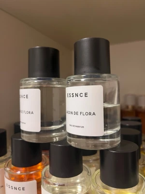 ESSNCE Jardin de Flora parfym - Obs! Den främre i bild! Se separat annons för den bakre! Minimalistisk parfymflaska från ESSNCE med doften Jardin de Flora. Flaskan är cylindrisk i genomskinligt glas med svart rund kork och vit etikett med svart text. Eau de Parfum-varianten ger en elegant och stilren känsla, perfekt för dig som gillar enkel design.