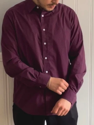 Mörklila/vinröd skjorta med knappar - Snygg mörklila/vinröd skjorta med klassisk krage och vita knappar framtill. Skjortan har lång ärm och rak passform, perfekt för dig som vill ha en stilren look. Tillverkad i mjuk bomull som känns skön mot huden.