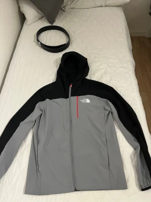Grå och svart vindjacka The North Face - Snygg vindjacka från The North Face i grått och svart med huva och hel dragkedja. Jackan har en röd dragkedja på bröstfickan och logga tryckt i vitt på bröstet. Tillverkad i lätt och tåligt material, perfekt för blåsiga dagar.