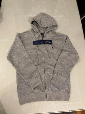 Grå zip hoodie från Polo Ralph Lauren - Klassisk grå zip hoodie från Polo Ralph Lauren med broderad logga på bröstet. Hoodien har dragsko i huvan, två fickor framtill och ribbade muddar vid ärmar och nederkant. Tillverkad i mjukt bomullsmaterial för en skön och avslappnad look.
