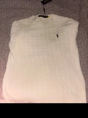 Stickad Polo tröja - Kabelstickad vit tröja från Polo Ralph Lauren med rund halsringning och den klassiska broderade loggan på bröstet. Tröjan har långa ärmar och ribbade muddar vid ärmslut och nederkant. Perfekt för en clean och stilren look. 