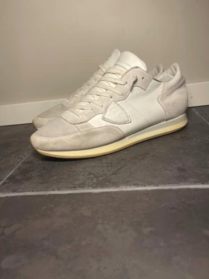 Vita sneakers från Philippe Model - Stilrena vita sneakers från Philippe Model med ovandel i mocka och textil, samt detaljer i läder. Klassisk låg modell med vita skosnören och beige sula. Perfekta för dig som gillar minimalistisk och clean stil.