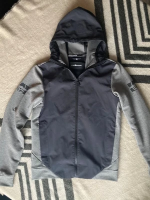 Grå hoodiejacka från Sail Racing - Snygg grå hoodiejacka från Sail Racing med dragkedja framtill och huva. Jackan har mörkare grå front och huva, ljusare grå ärmar samt logotypdetaljer på båda ärmarna. Tillverkad i ett mjukt och stretchigt material som känns sportigt och modernt.