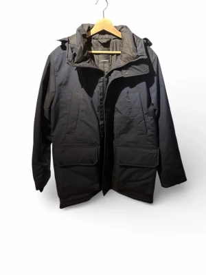 Marinblå J.Lindeberg vinterjacka med huva - Snygg marinblå vinterjacka med huva och stora framfickor. Jackan har dragkedja och knappslå framtill samt justerbar huva. Perfekt för kalla dagar och har en klassisk, rak passform. Fodrad insida för extra värme.