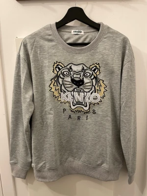 Grå Kenzo sweatshirt - Säljer en grå sweatshirt från Kenzo med det ikoniska tigerhuvudet broderat framtill och texten 'KENZO PARIS' under. Tröjan har rund halsringning, ribbade muddar och är gjord i mjukt bomullsmaterial. Perfekt för dig som gillar streetwear och vill sticka ut.