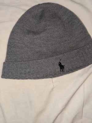 Grå mössa från Polo Ralph Lauren - Snygg grå mössa från Polo Ralph Lauren i mjuk merinoull. Klassisk ribbad kant och broderad logga framtill. Perfekt för dig som vill ha en stilren och bekväm accessoar till hösten och vintern.