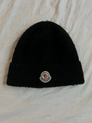 Moncler mössa  - Svart ribbstickad mössa från Moncler med uppvikt kant och klassisk Moncler-logga framtill. Tjockt garn ger en chunky känsla och mössan har en enkel, stilren design som passar till det mesta. Perfekt för dig som gillar streetwear och exklusiva märken.