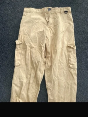 Beige cargobyxor med fickor - Säljer ett par beige cargobyxor med klassiska stora sidofickor och rak passform. Byxorna har bälteshällor och stängs med knapp och dragkedja. Perfekta för en avslappnad och trendig stil. Materialet ser ut att vara bomull eller bomullsblandning.