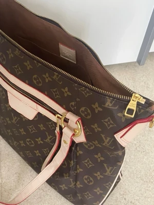 Louis Vuitton Monogram handväska - Säljer en klassisk Louis Vuitton handväska i brunt monogramcanvas med beige läderdetaljer och röda kanter. Väskan har guldiga metalldetaljer, dragkedja upptill och en praktisk ytterficka med dragkedja. Perfekt för dig som gillar lyxig och tidlös design.