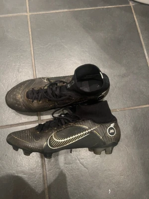 Nike Mercurial Superfly fotbollsskor -  svarta Nike Mercurial Superfly fotbollsskor med guldfärgade detaljer och vit swoosh. Skorna har strumpliknande skaft, snörning och dobbar som passar överallt. Tillverkade i syntetmaterial för bra grepp och kontroll på planen.