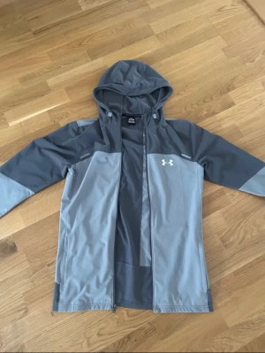 Grå vindjacka från Under Armour - Snygg grå vindjacka från Under Armour med huva och dragkedja. Jackan har två nyanser av grått, logga på bröstet och stort Under Armour-tryck på ryggen. Tillverkad i lätt och tåligt syntetmaterial, perfekt för blåsiga dagar. Jacka har en pytte defekt vid dragkedja!