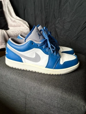 Nike Air Jordan 1 Low blå/vit - Snygga Nike Air Jordan 1 Low sneakers i blått och vitt läder med grå detaljer. Klassisk siluett med perforerad tåbox, blå snörning och ikoniska Jumpman-loggan på plösen. Perfekta för dig som vill ha en stilren och sportig look. Sitter mer som 41or