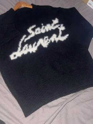 Svart Saint Laurent stickad tröja - Säljer en svart stickad tröja från Saint Laurent med vit fuzzy logga framtill. Tröjan har rund halsringning och ribbade muddar vid ärmslut och nederkant. Perfekt för dig som vill ha en statement piece i garderoben.