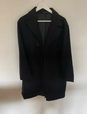 Svart klassisk ullkappa, Tiger of Sweden ”Fredde” - Stilren svart ullkappa med tre knappar framtill och breda slag. Kappan har rak passform och två sidofickor. Storlek 48, från Tiger of Sweden i modellen ”Fredde”. Mörkgrå/svart. Nypris 4999kr