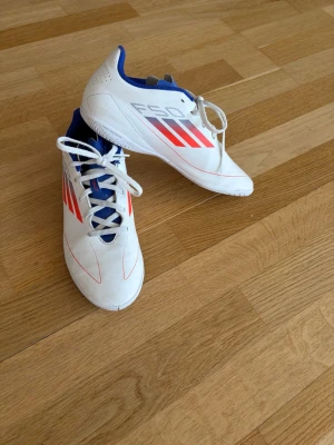 Vita Adidas fotbollsskor med röd detalj - Snygga vita fotbollsskor från Adidas med röda och silverfärgade detaljer samt blå insida. Skorna har klassisk snörning och låg profil, perfekta för snabba vändningar på planen. Tillverkade i syntetmaterial för bra passform och lätt känsla.