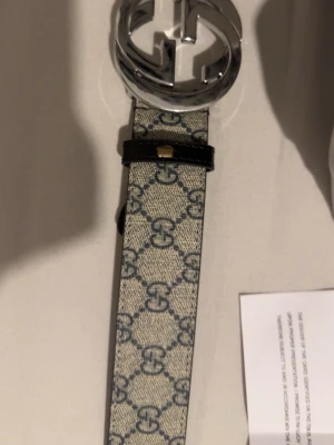 Gucci beige bälte med GG-mönster - Säljer ett snyggt beige bälte från Gucci med klassiskt GG-mönster i blått och en stor silverfärgad GG-spänne. Bältet är tillverkat i canvas med detaljer i svart skinn. Perfekt för att lyfta din outfit med en lyxig touch.