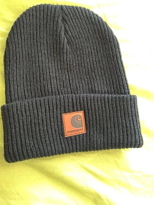 Svart ribbstickad mössa Carhartt - Svart ribbstickad mössa från Carhartt med uppvikt kant och klassisk läderpatch med logga framtill. Snygg och enkel design som passar till det mesta. Tillverkad i mjukt och stretchigt material för skön passform.