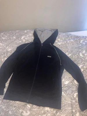 Svart zip hoodie från BOSS - Snygg svart hoodie från BOSS med dragkedja och vit piping längs axlarna. Hoodien har en grå insida i huvan och BOSS-logga broderad på bröstet. Perfekt för en chill och stilren look. pris kan diskuteras!