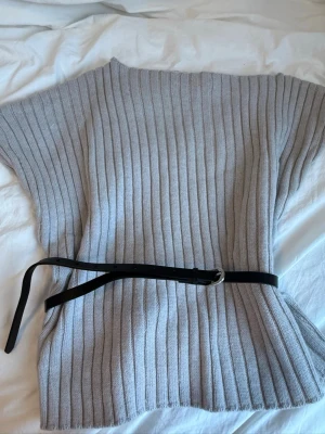 Grå ribbstickad väst med bälte - Säljer en grå ribbstickad väst med korta ärmar och rund hals. Västen har ett svart smalt bälte i midjan som ger en snygg siluett. Perfekt att styla över en långärmad tröja eller skjorta. Materialet känns mjukt och stretchigt.