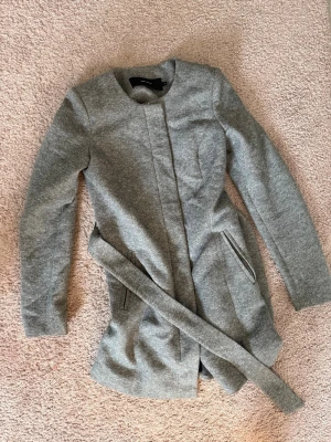 Grå kappa från Vero Moda med bälte - Stilren grå kappa från Vero Moda i storlek XS. Kappan har ett avtagbart bälte i midjan, två snedställda fickor med dragkedja och rund halsringning. Materialet är mjukt och har en lätt melerad look. Perfekt för dig som gillar minimalistisk stil.