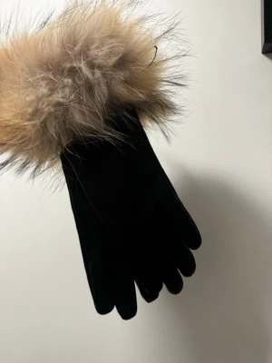 Svarta skinnhandskar med pälsdetalj - Stilrena svarta handskar i skin med bred, fluffig pälsdetalj i beige och bruna toner vid handleden. Perfekta för dig som vill ha en lyxig accessoar till kallare dagar. Handskarna har en klassisk form och känns riktigt mjuka.