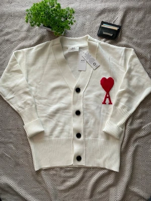 Vit kofta från Ami Paris med rött hjärta - Tja! Jag aäljer en Vit kofta från Ami Paris med svart knappstängning och ribbade muddar. På bröstet finns ett stort rött hjärta med bokstaven A under. Koftan har en avslappnad passform och är tillverkad i mjukt stickat material. Perfekt för dig som gillar stilrena plagg med en twist.