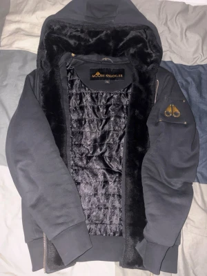 Svart hoodie från Moose Knuckles - Snygg svart hoodie från Moose Knuckles med dragkedja, huva och mjukt foder i både kropp och huva. Har en guldfärgad logga på ärmen och ribbade muddar. Perfekt för dig som vill ha en varm och stilren hoodie med exklusiv känsla. PRISET KAN DISKUTERAS!!!