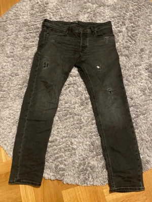Svarta slitna skinny jeans - Säljer ett par svarta jeans tapered Mike med slitningar och hål på benen för en trendig och avslappnad look. Jeansen har klassisk femficksmodell och dragkedjegylf. Perfekta för dig som gillar streetstyle och vill ha ett par jeans med attityd. Storlek 32/32 