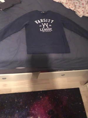 Svart sweatshirt Warp Varsity League - Cool svart sweatshirt från Warp med trycket 'Varsity League 1996' i vitt framtill. Tröjan har rund halsringning, långa ärmar och en ficka med dragkedja på sidan. Perfekt för en avslappnad och sportig stil.