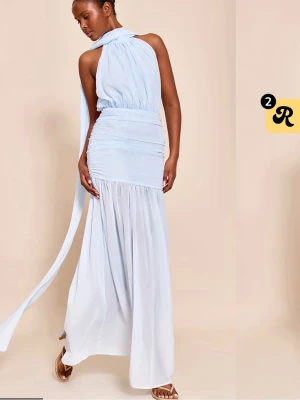 Ljusblå långklänning med omlottkrage - Elegant ljusblå långklänning med omlottkrage och långa band som faller ner längs ryggen. Klänningen har markerad midja, rynkade detaljer och är ärmlös med öppen rygg. Perfekt för sommarens tillfällen.