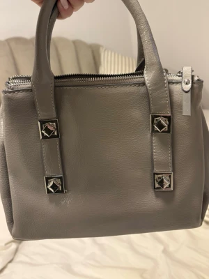 Grå handväska med nitar och dragkedjor - Snygg grå handväska i fejkläder med två handtag och coola silverfärgade nitar på framsidan. Väskan har flera fack med dragkedjor och svart innerfoder. Perfekt för dig som gillar stilrena detaljer och vill ha plats för allt viktigt.