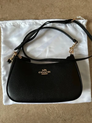 Svart axelremsväska från Coach - Stilren svart handväska från Coach i slätt läder med guldfärgade metalldetaljer. Väskan har justerbar axelrem och klassisk Coach-logga framtill. Perfekt storlek för det viktigaste och passar till många olika looks.