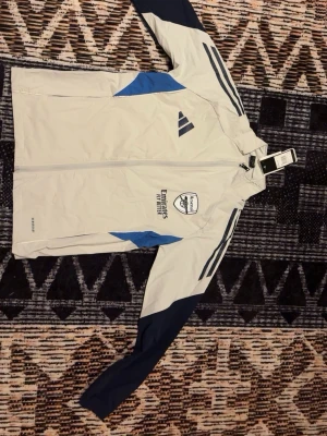 Vit Adidas Arsenal träningsjacka - Snygg vit träningsjacka från Adidas med Arsenal-logga och Emirates sponsortryck på bröstet. Jackan har blå detaljer och mörkblå ärmar samt klassiska Adidas-ränder på axlarna. Perfekt för fotbollsträning eller som sportig streetwear.