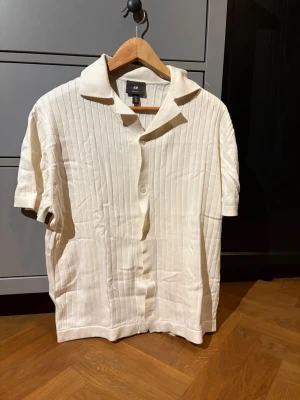 Beige ribbad skjorta  - Kortärmad beige skjorta från H&M med ribbad struktur och klassisk krage. Den är i storleken M och har en lite missfärgning på bild tre. Inga andra missfärgningar eller defekter som är synliga.