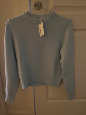 Ljusblå stickad tröja från H&M - En stilren ljusblå aldrig använd stickad tröja från H&M med rund halsringning och ribbade muddar vid ärmslut och nederkant. Tröjan har en enkel, klassisk design och är tillverkad i mjukt material som passar perfekt till vardagslooken.