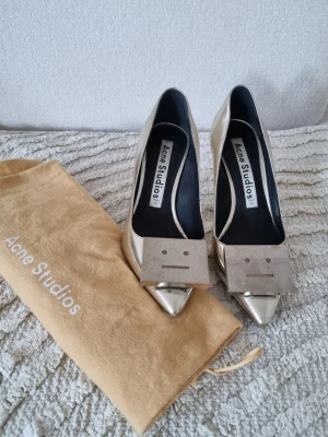 Silvriga pumps från Acne Studios - Säljer ett par silvriga pumps från Acne Studios med spetsig tå och hög klack. Skorna har en metallisk finish och en unik dekorativ detalj framtill med Acne Studios ikoniska ansikte. Kommer med original dustbag.