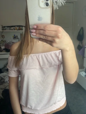 Ljusrosa off shoulder topp - Söt ljusrosa off shoulder topp med volangdetalj längs halsringningen och resår i nederkant. Kort ärm och croppad modell som ger en avslappnad och trendig look. Perfekt till jeans eller kjol för en fräsch sommarstil. Den går att strykas. Passar xxs/xs/s