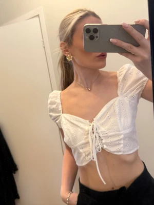 Vit croppad blus med snörning - Superfin vit croppad blus med puffärmar och broderade detaljer. Toppen har snörning framtill och v-ringad hals. Perfekt för sommaren och ger en romantisk vibe till din outfit.