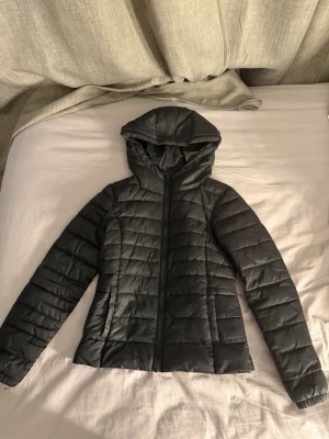 Svart pufferjacka med huva - Snygg svart pufferjacka med huva och dragkedja framtill. Jackan har quiltad design med horisontella sömmar och två sidofickor. Perfekt nu inför våren och enkel att matcha med olika outfits. Materialet är troligtvis syntet, vilket gör den lätt och varm.