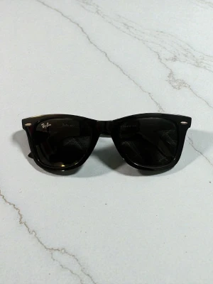 Svarta Ray-Ban Wayfarer solglasögon - Klassiska svarta Wayfarer solglasögon från Ray-Ban med tidlös design och mörka glas. Bågarna är i blank svart plast och har Ray-Ban-loggan på sidan. Perfekta för dig som vill ha en ikonisk och stilren look.