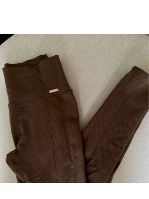 Bruna ribbade leggings Aim'n - Säljer ett par bruna leggings från Aim'n med hög midja och ribbad struktur. De är stretchiga och sitter tight, perfekta för både träning och chill. Metallogo på höften ger en snygg detalj. Materialet är mjukt och följsamt.