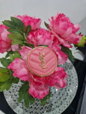 YSL rosa puderrouge - Lyxigt puderrouge från Yves Saint Laurent i en rund, guldig dosa med quiltad rosa ovansida och YSL-logga i guld. Färgen är en mjuk rosa ton som ger kinderna en fräsch och naturlig look. Perfekt för att addera glow till din makeup.