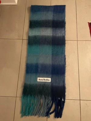Rutig blågrön halsduk Acne Studios - Säljer en fluffig halsduk från Acne Studios i rutigt mönster med olika nyanser av blått, grönt och turkos. Halsduken har långa fransar i ändarna och en stor Acne Studios-logga. Perfekt för att addera färg till din outfit.