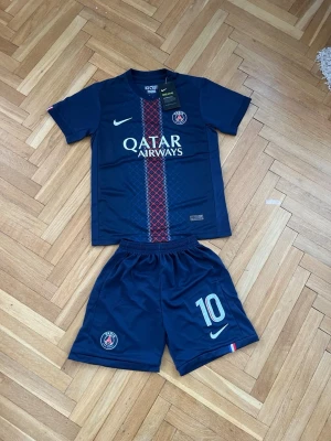 PSG matchställ O. Dembélé #10 Nike - Mörkblått PSG matchställ med tröja och shorts. Tröjan har Qatar Airways-tryck, PSG-logga och Nike-märke samt rödblått mönster på framsidan. Nummer 10 och O. Dembélé på ryggen. Shortsen har PSG-logga och nummer 10. Tillverkat i lätt funktionsmaterial.