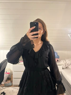 Svart volangklänning med v-ringning från bubbleroom  - Snygg svart kortklänning med v-ringning och volangdetaljer. Klänningen har långa, transparenta ärmar och markerad midja. Använd vid endast 1 tillfälle dock som man kan se på sista bilderna har de blivit lite trådar som lossnat😔 men inget som förstör helhetsintrycket! Nypris över 600 
